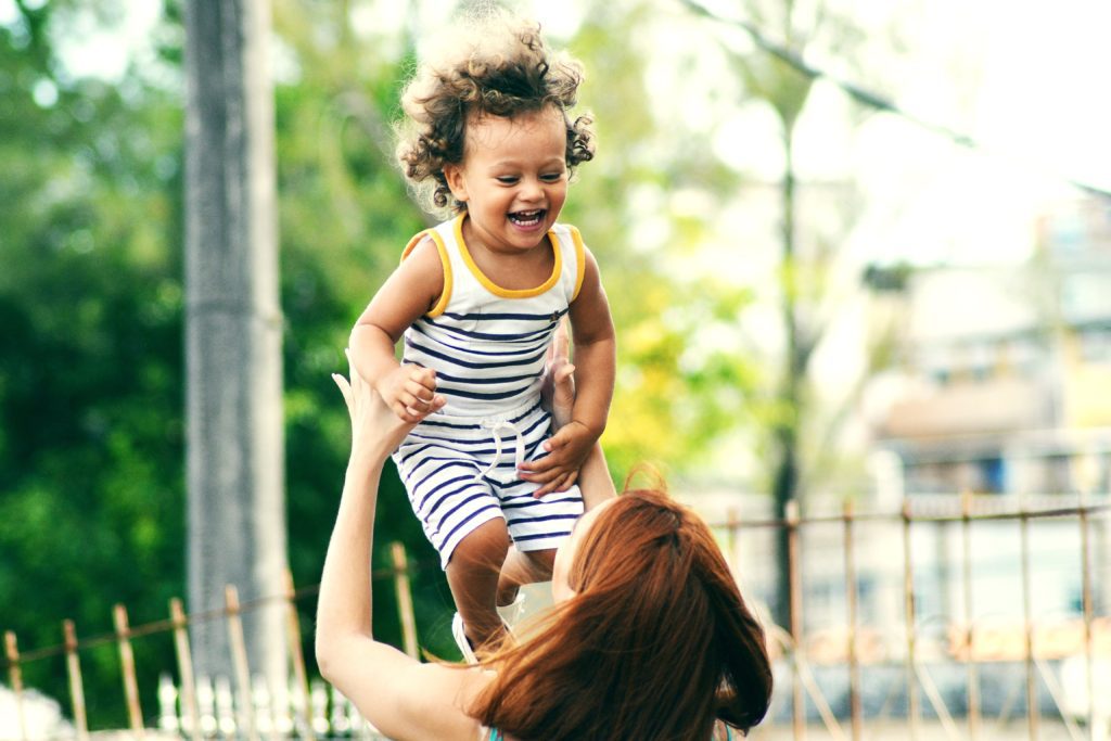 Young, curly haired boy in striped playsuit being tossed airborn by red-headed mother in a neighborhood park