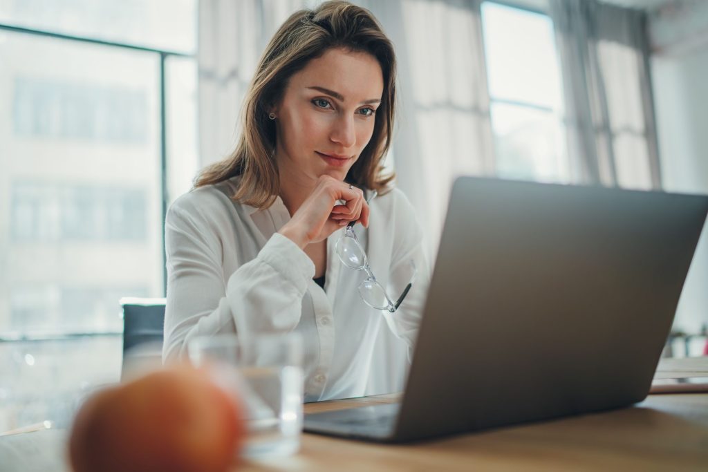 Female paralegal sits at a desk studying information on her laptop while holding her glasses.